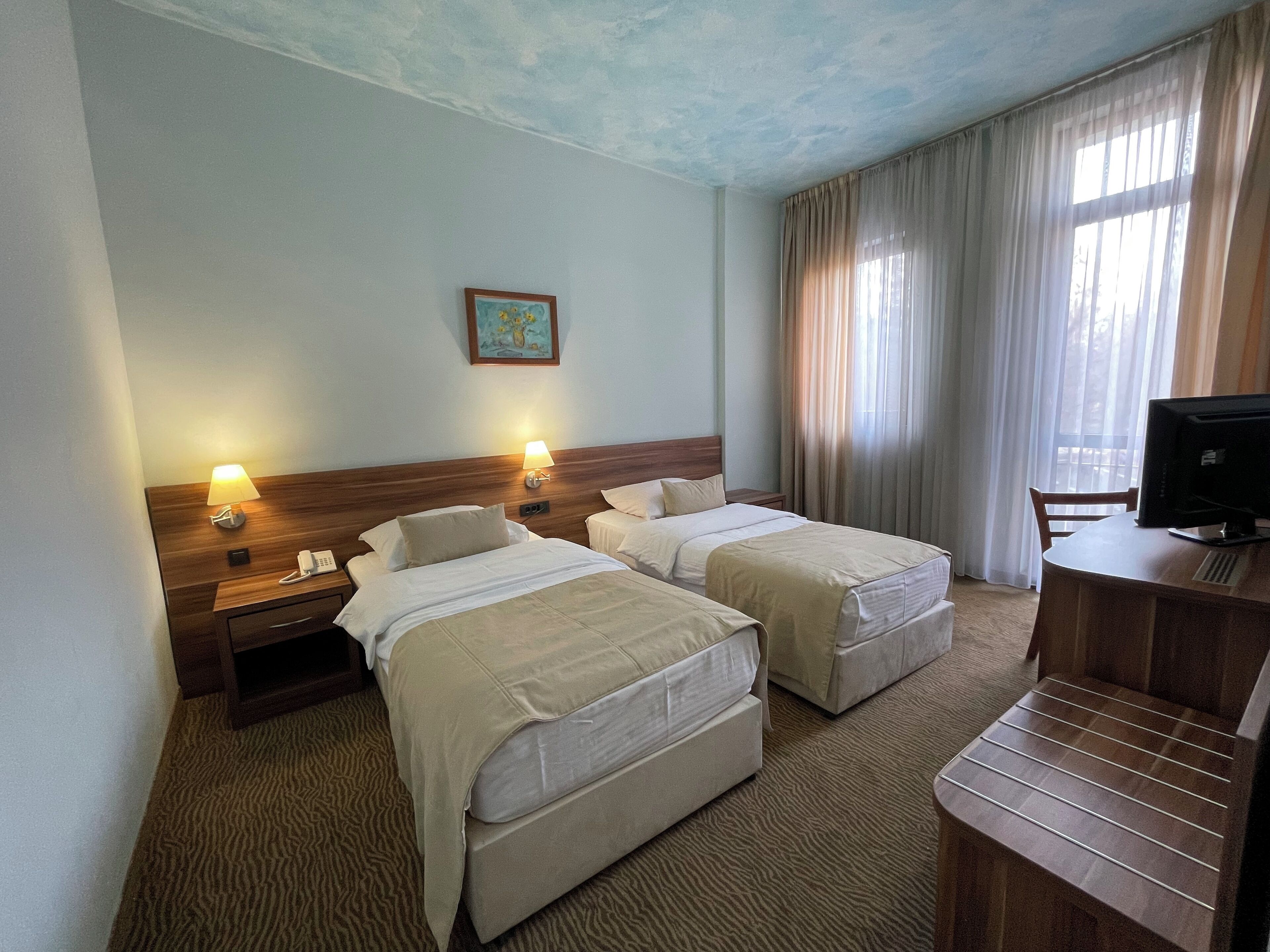 A hotel room with two beds, a desk with a computer, a telephone, and a painting on the wall.