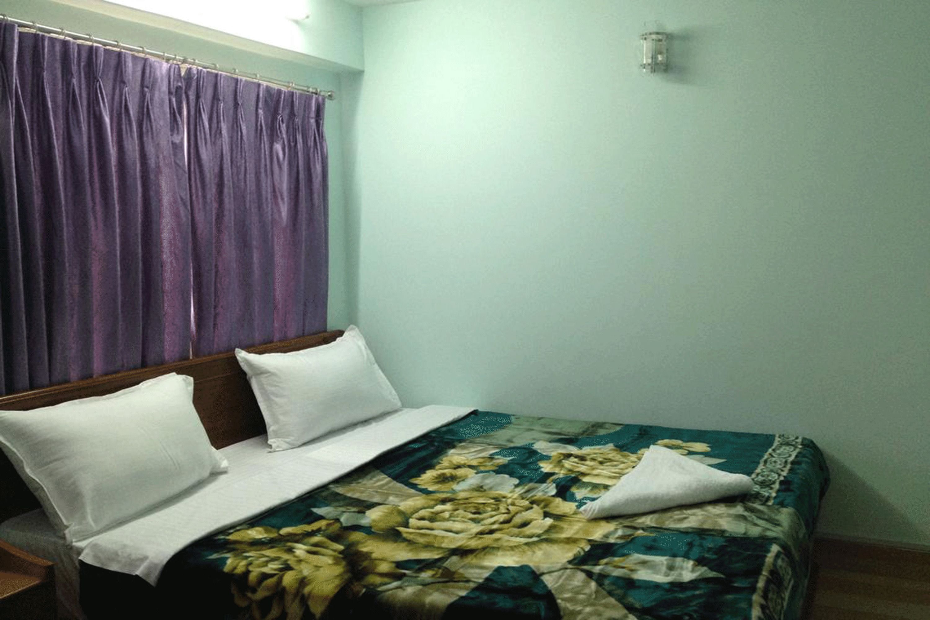 A single bed with white pillows and a floral bedspread in a room with light green walls and a purple curtain.