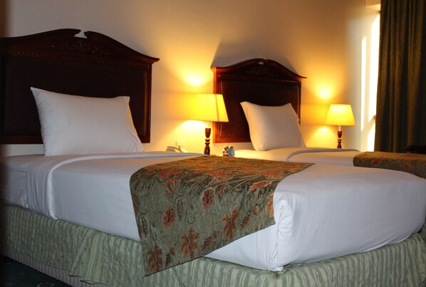 A hotel room with two beds, each with a wooden headboard, white bedding, and a green patterned throw. There are two bedside lamps and a nightstand with a lamp. - Mount Royal Hotel (Dubai)