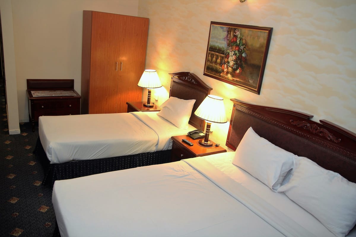 A hotel room with two single beds, a wooden wardrobe, a nightstand with a lamp, and a painting on the wall.
