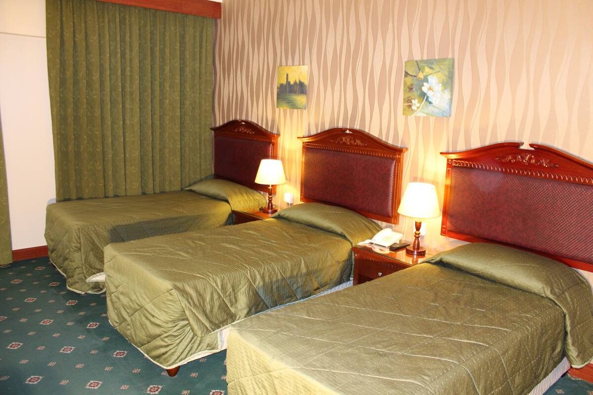 A hotel room with three single beds, each with a wooden headboard and a green bedspread. There are two bedside tables with lamps, and the room has a patterned carpet.