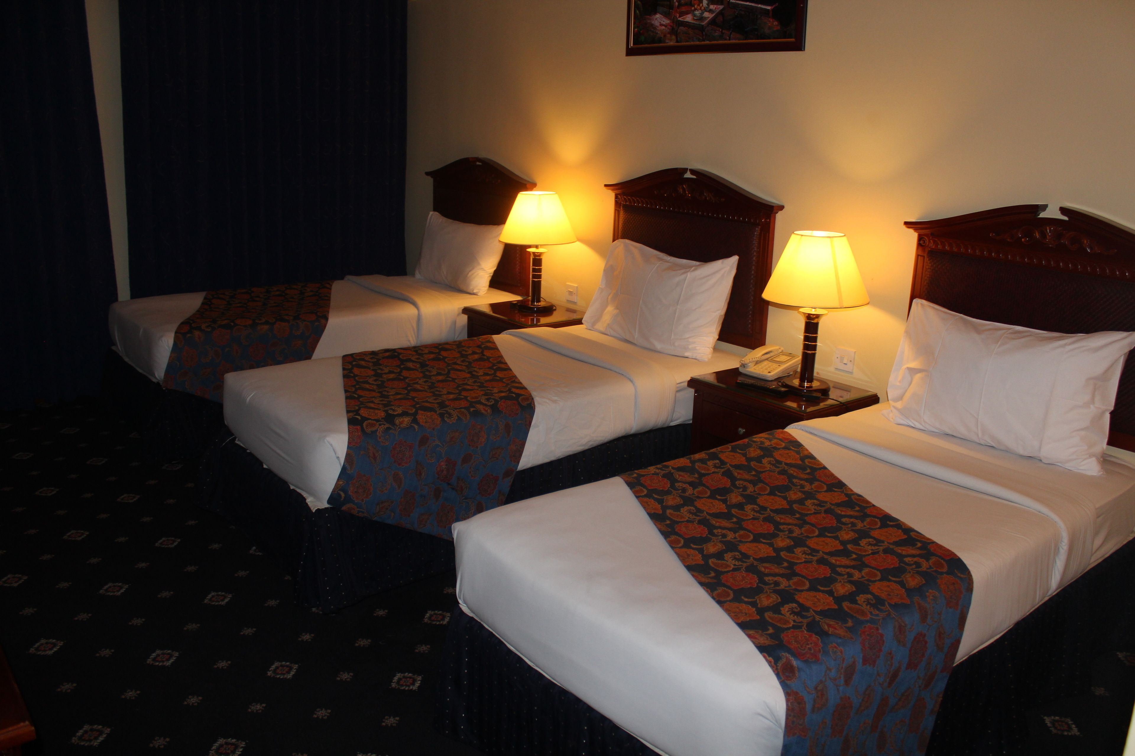 A hotel room with three beds, each with a patterned bedspread and white pillows. There are bedside tables with lamps and a telephone.