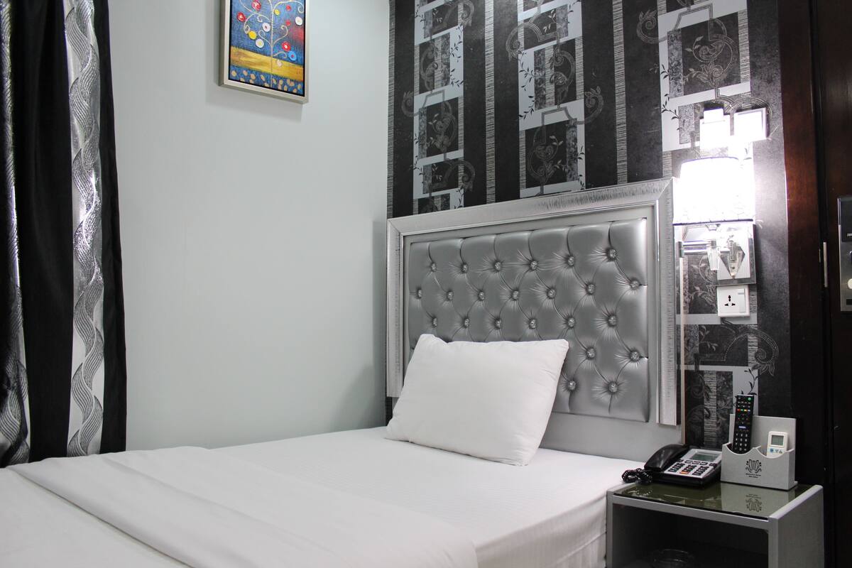 A neatly made bed with a white pillow and a tufted headboard, a bedside table with a phone and a lamp, and a wall with a patterned wallpaper and a framed picture.