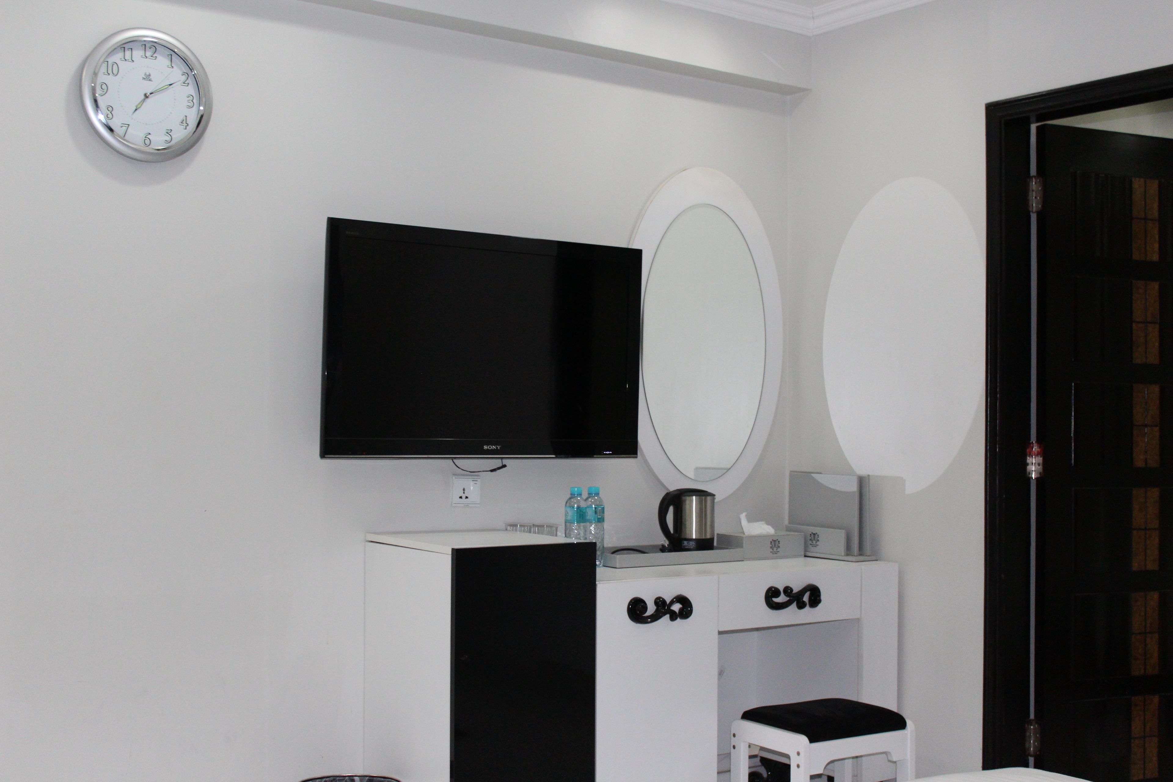 A room with a flat-screen TV, a white vanity with a mirror, a small stool, and a clock on the wall.