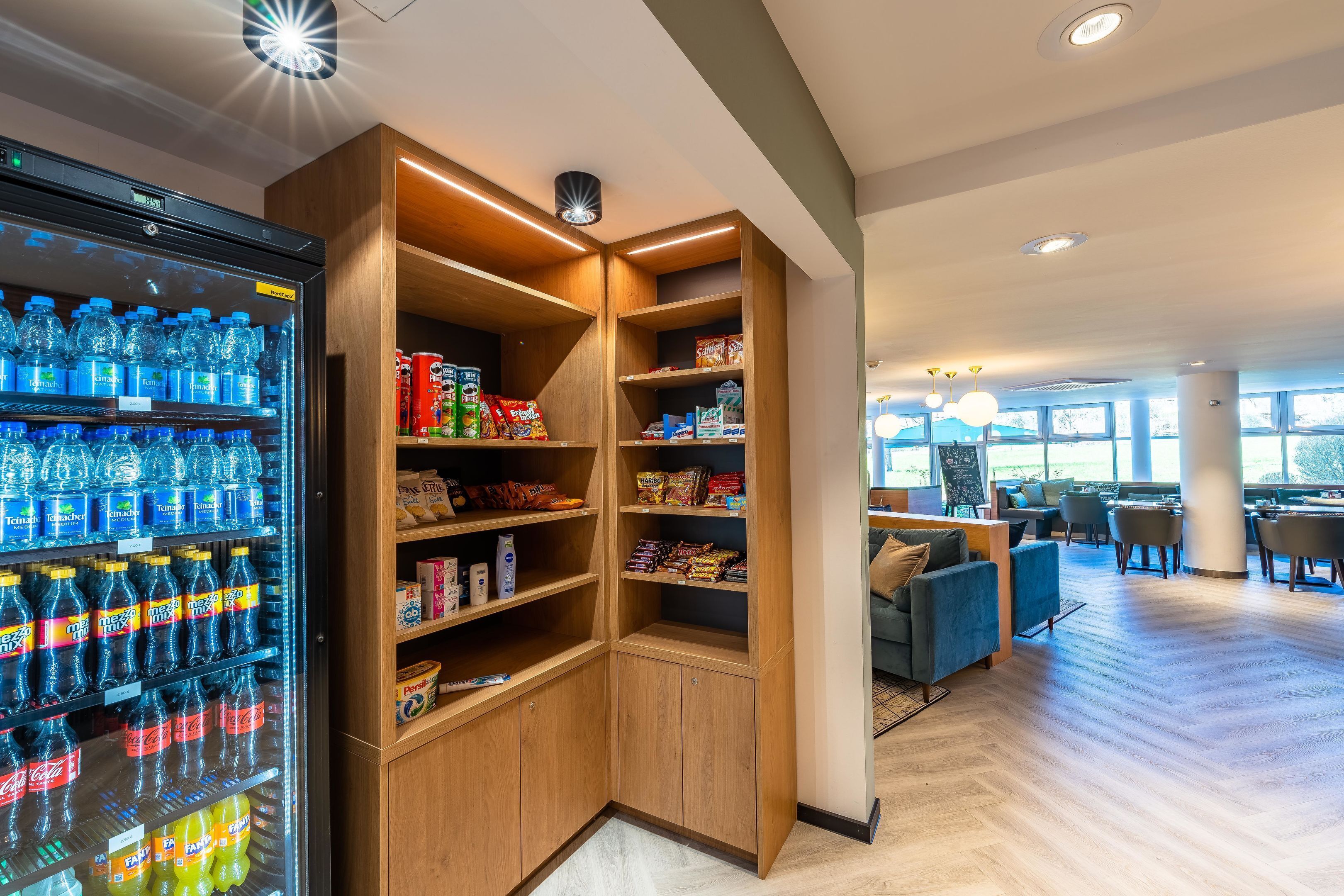 Eine Ecke der Hotelhalle mit einem Getränkekühlschrank, gefüllt mit verschiedenen Getränken, Holzregalen mit Snacks und Toilettenartikeln sowie einer Sitzecke mit Tischen und Stühlen.