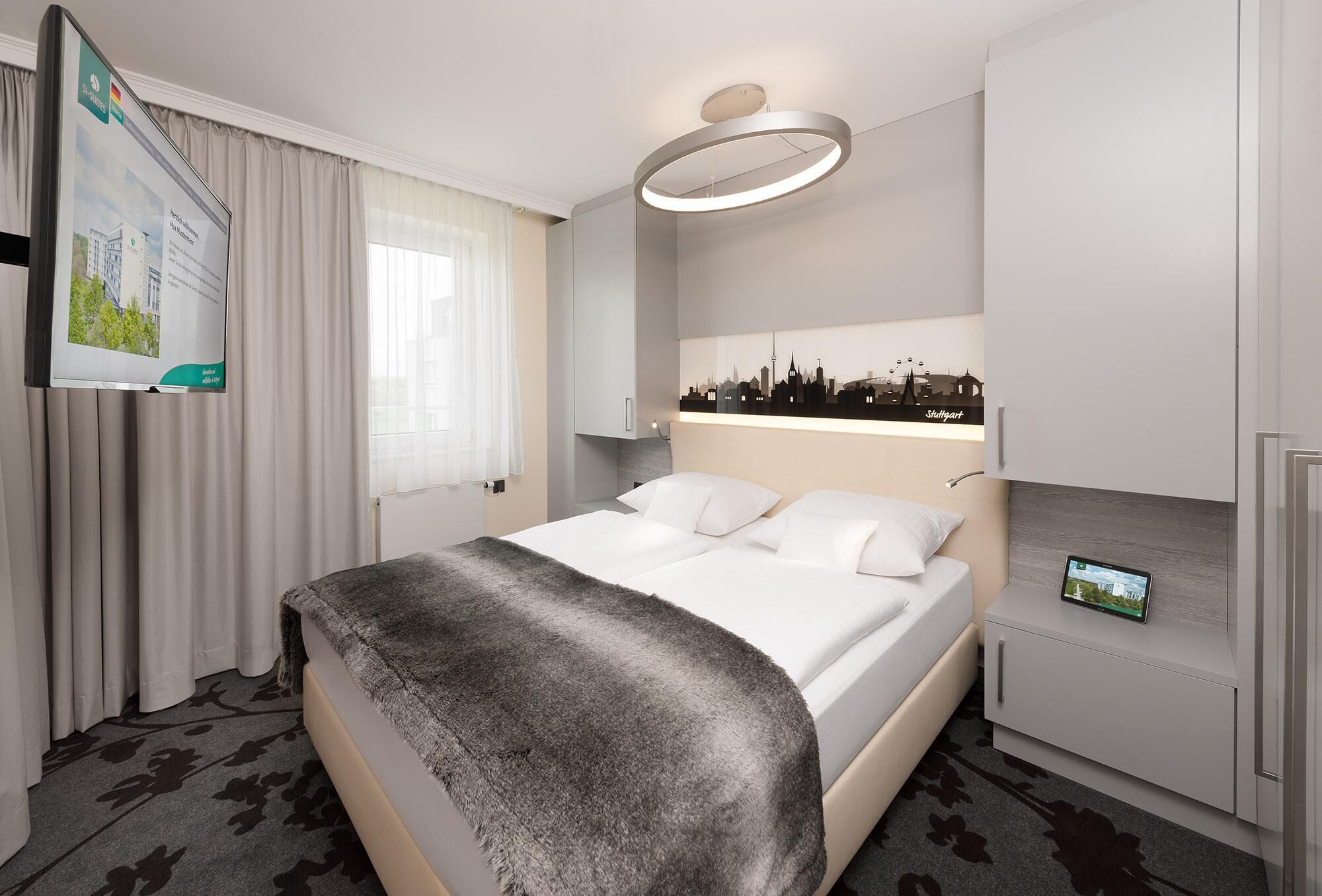 Camera d'albergo moderna con un letto, uno schermo digitale, un'opera d'arte con una veduta cittadina e un armadio a muro.