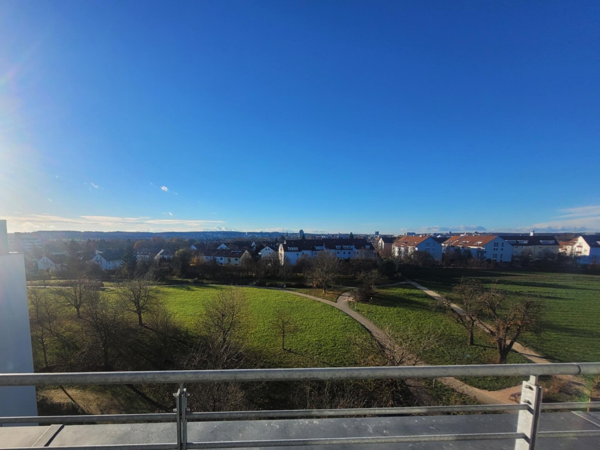 Blick vom Balkon auf ein Wohngebiet mit Häusern, grünen Feldern und blauem Himmel.