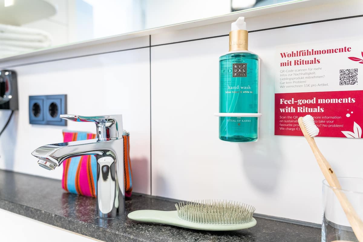 A bathroom with a modern faucet, a toothbrush holder with colorful towels, a hand wash bottle, and a promotional sign.