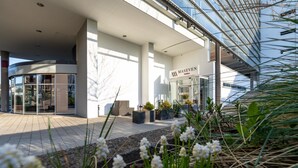L’entrée moderne de l’hôtel se distingue par sa façade en verre, une enseigne indiquant « MASEVEN Suites » et un espace extérieur aménagé avec des plantes.