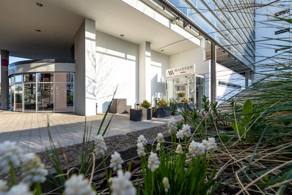 A modern hotel entrance with a glass facade, a sign reading "MASEVEN Suites," and a paved outdoor area with plants.
