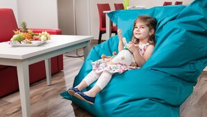 Un enfant est assis sur un fauteuil en forme de poire de couleur turquoise dans une pièce où se trouvent une table blanche, des chaises rouges et un vase rempli de fleurs.