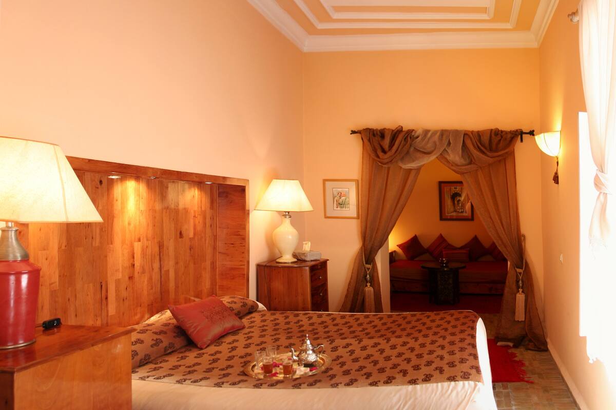 A hotel room with a wooden headboard, a bed with patterned bedding, a bedside table with a lamp, a nightstand with a lamp, a red rug, and a wall-mounted picture.
