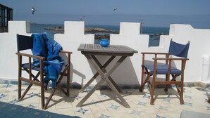 A terrace with two wooden chairs and a table, overlooking a coastal view with rocks and the sea. - Dar Loulema (Essaouira)