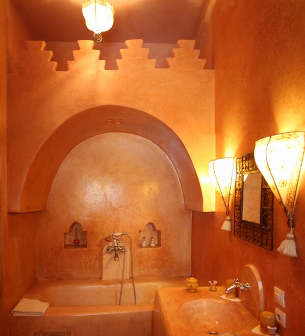 Un baño con bañera, lavamanos y espejo. Hay dos luces empotradas en la pared y un arco decorativo sobre la bañera.