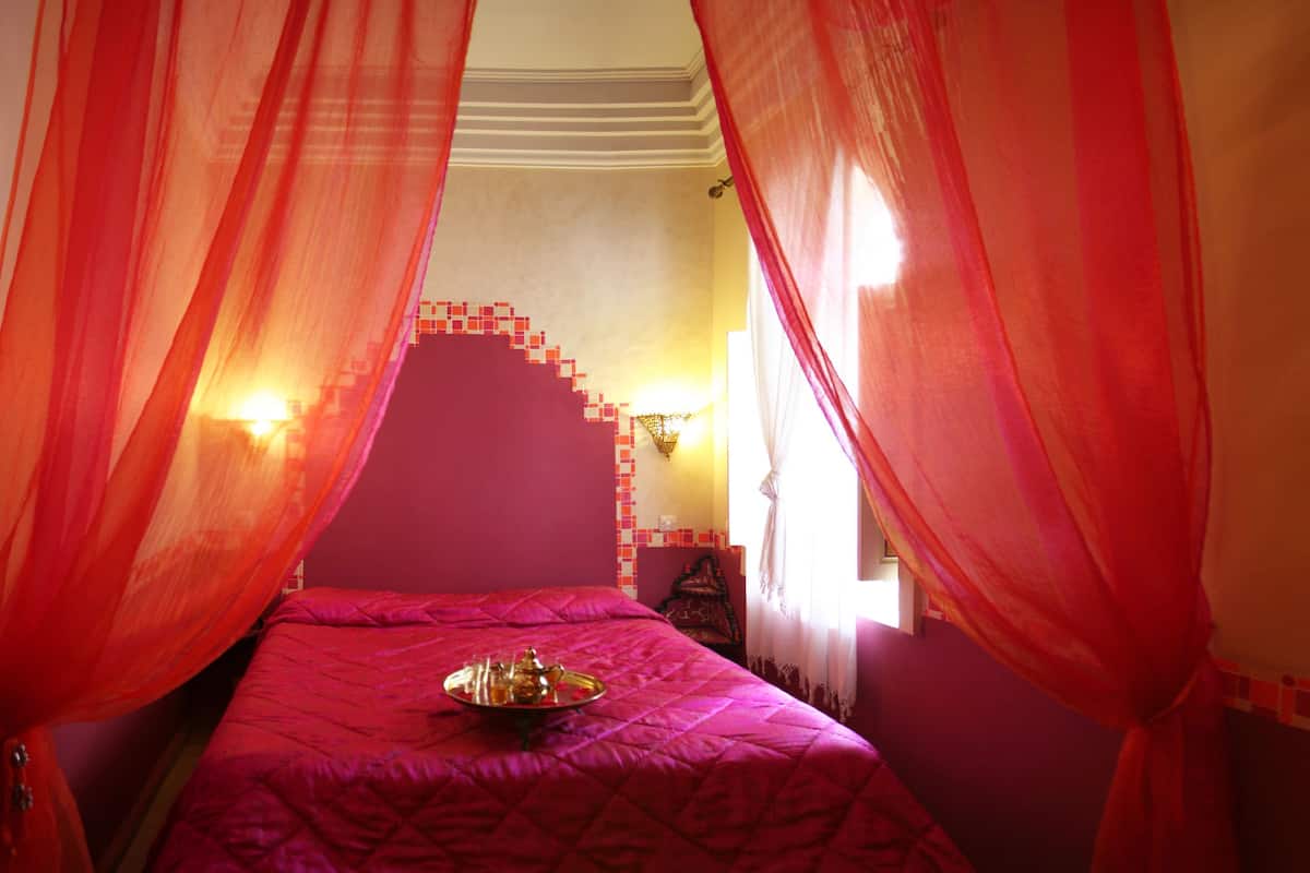 A room with a bed covered in a red bedspread, a tray of items on the bed, and a doorway leading to another room with a tiled floor and a vase with flowers.