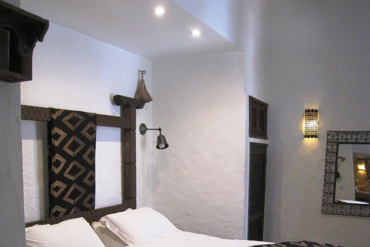 A neatly made bed with patterned bedding, a wall-mounted light fixture, and a mirror with a decorative frame.