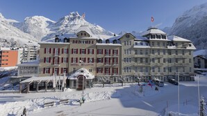 A hotel with multiple floors, a snow-covered landscape, and mountains in the background. - Bellevue-Terminus - Urban Lifestyle Hotel (Engelberg)