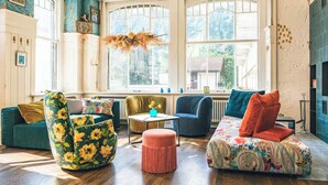 A cozy living room with a large window, a green floral armchair, a blue sofa, and a patterned ottoman. - Bellevue-Terminus - Urban Lifestyle Hotel (Engelberg)