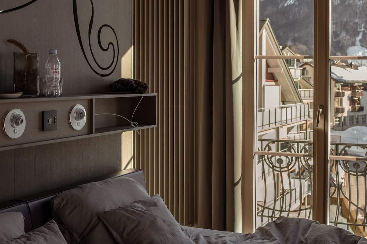 Un dormitorio con una cama, una estantería con objetos, un radiador y una ventana con vistas a edificios.