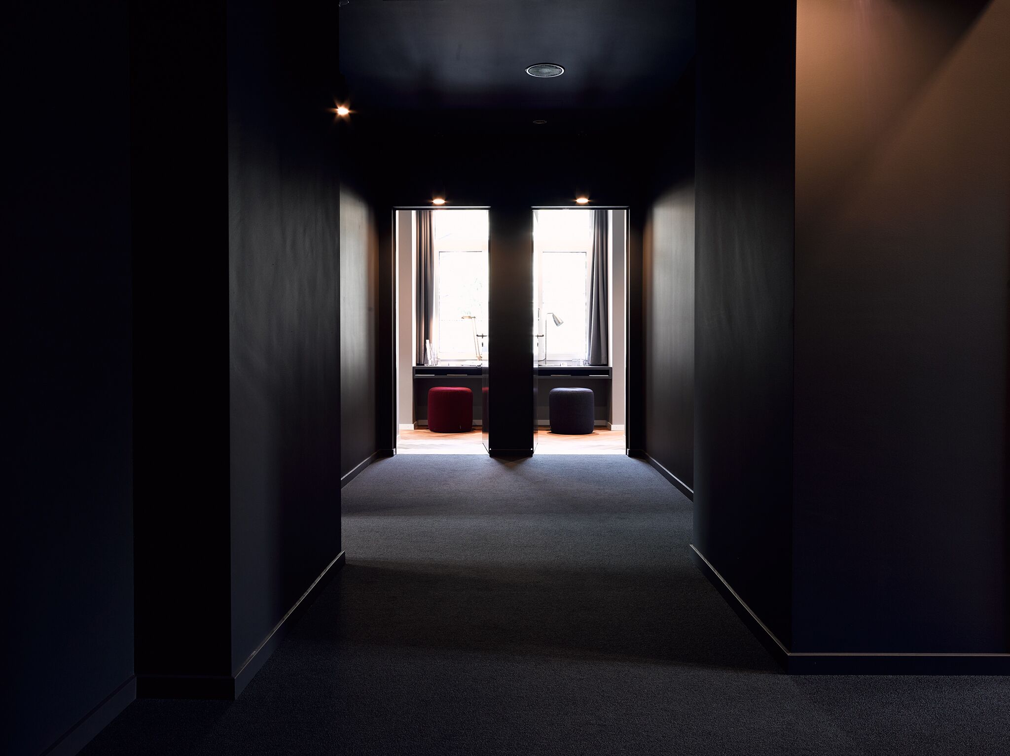 A narrow hallway with a carpeted floor leading to a room with a window and a desk.