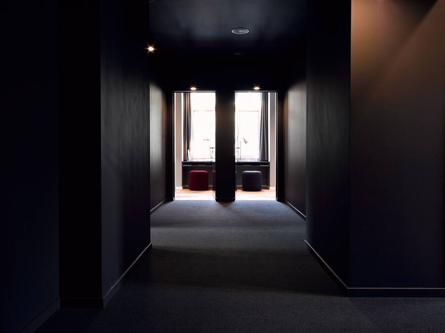 Un pasillo estrecho con piso alfombrado que conduce a una habitación con ventana y escritorio.