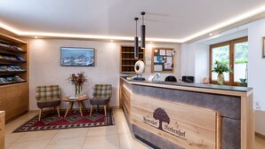 Un espace d’accueil comprenant un comptoir de réception en bois, deux chaises, une petite table avec un vase, un tableau accroché au mur et une fenêtre avec des stores.