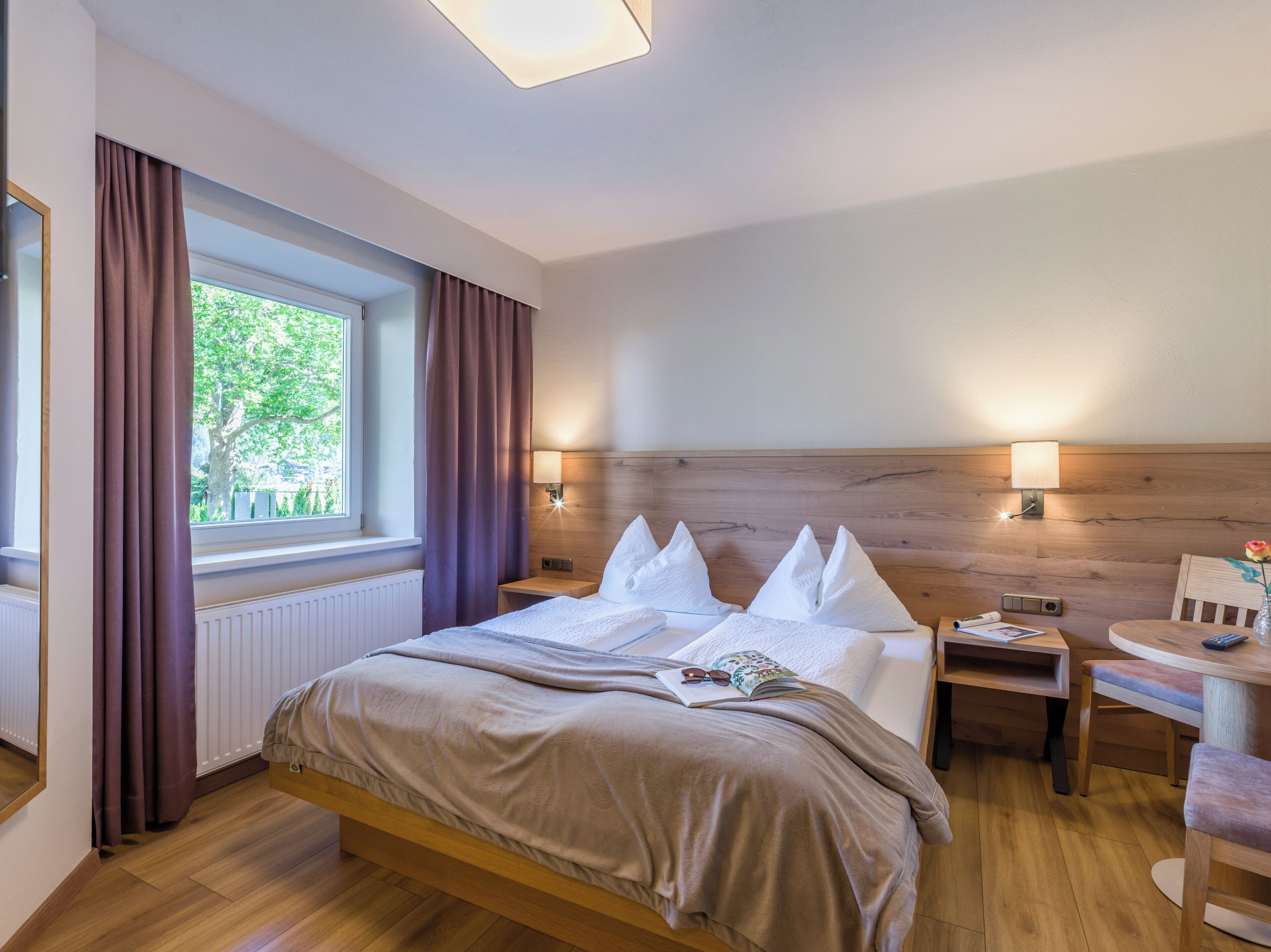 A neatly made bed with white linens, a bedside table with a lamp, and a window with curtains.