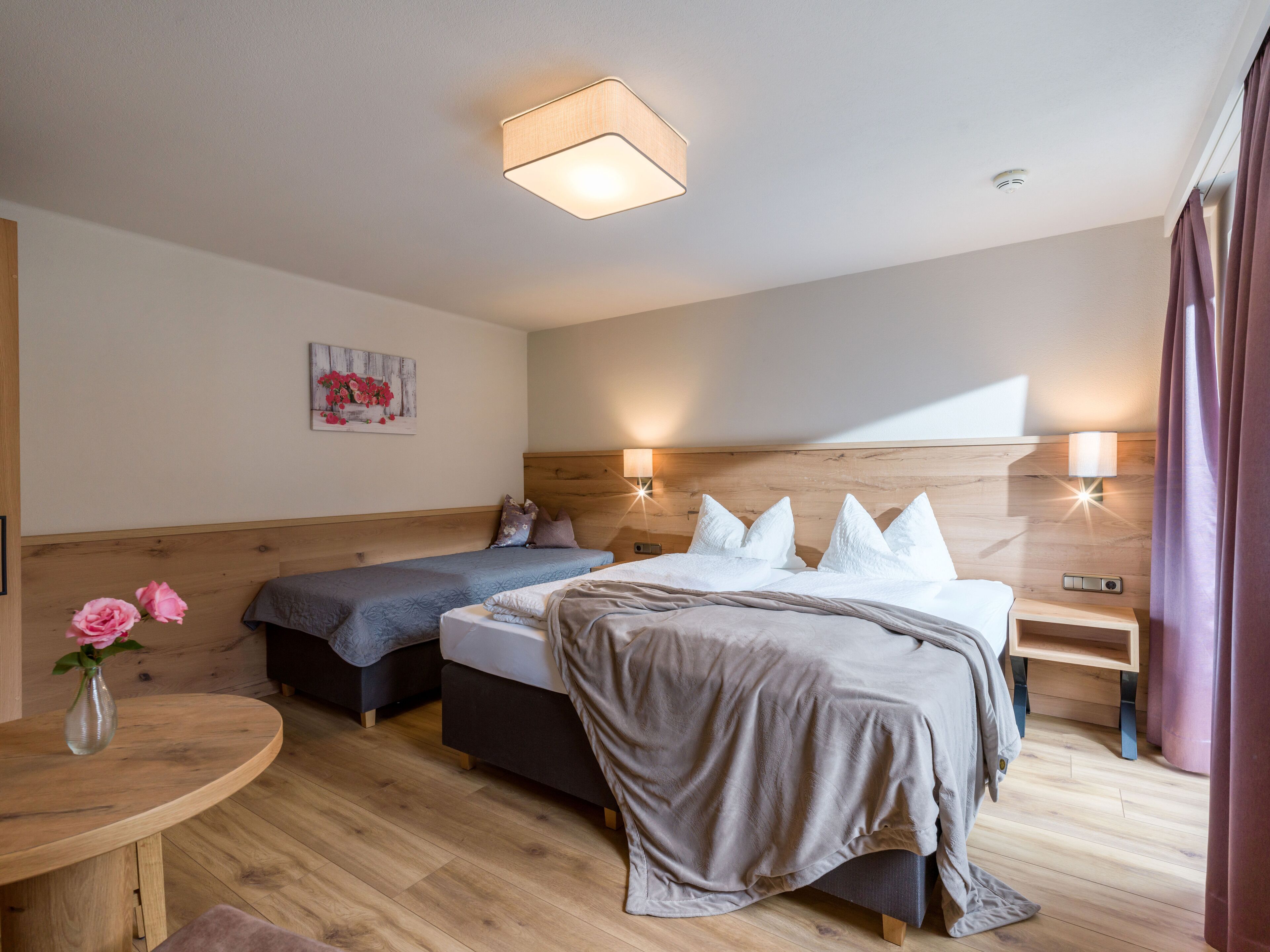 A hotel room with a wooden headboard, two beds, a bedside table, a nightstand, a small table with a vase of flowers, and a wall-mounted light fixture.