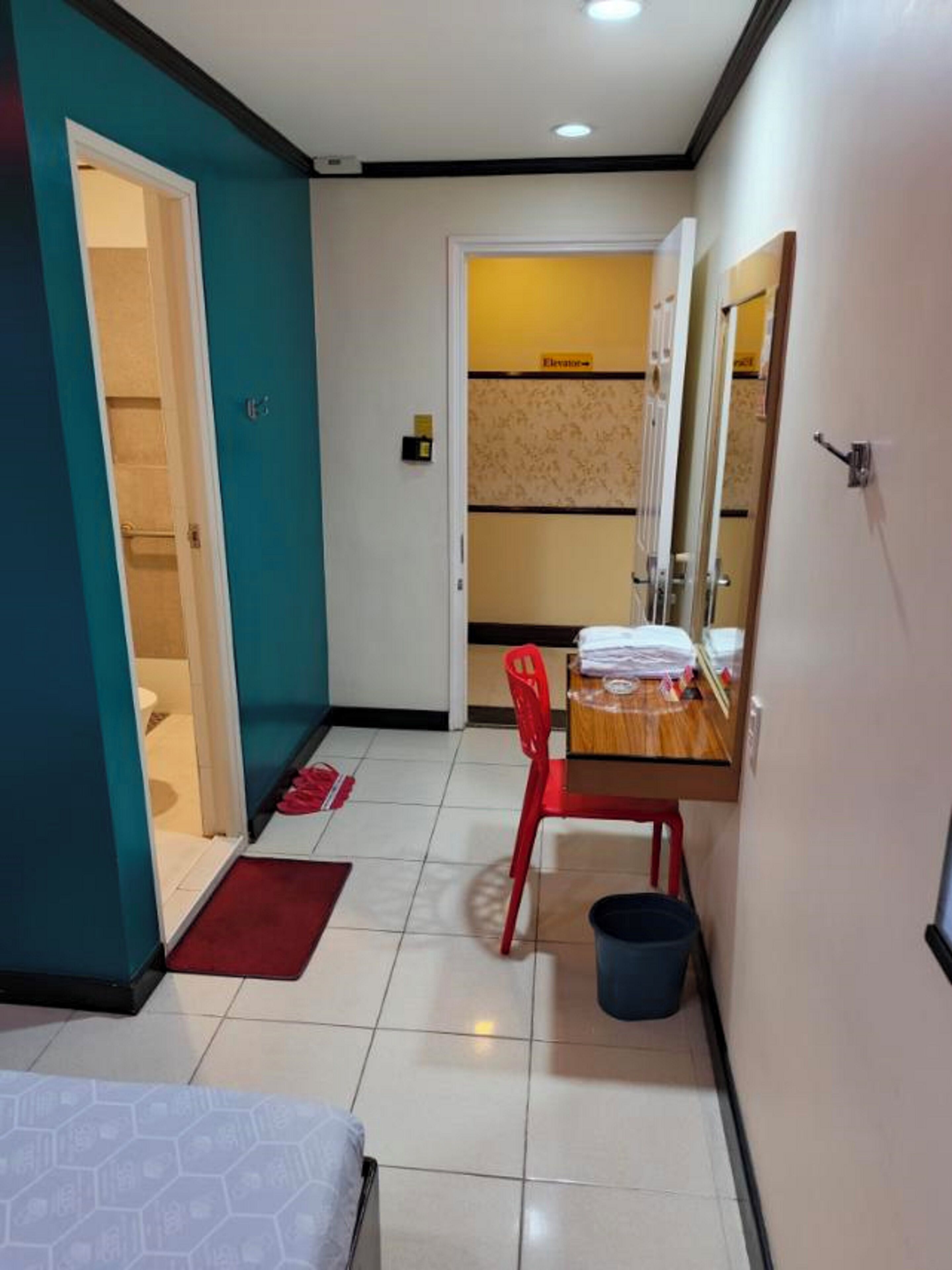 A hotel room with a bed, a desk with a chair, a red chair, a small trash bin, and a bathroom with a toilet and shower.