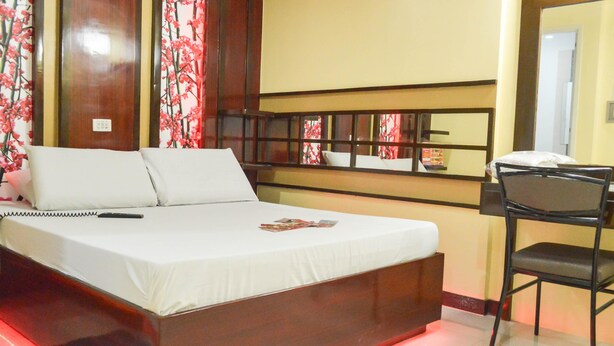A neatly made bed with white linens and pillows, a bedside table with a remote control, and a chair with a cushion.