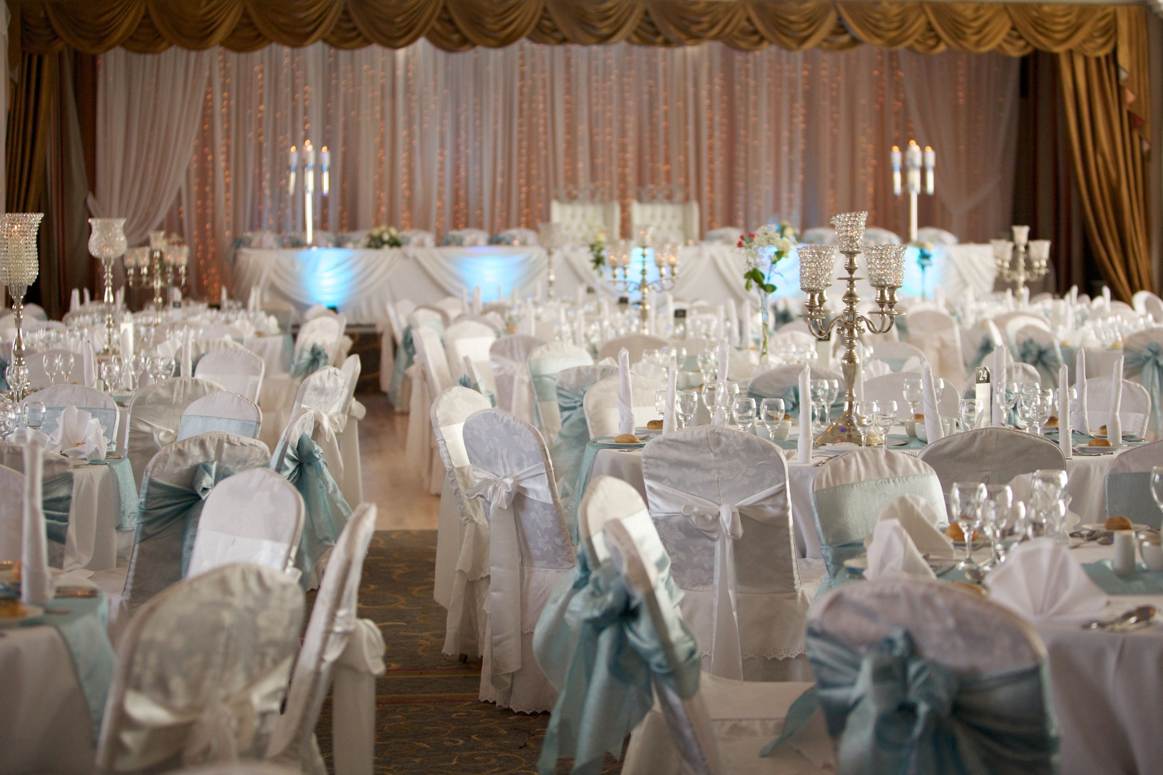 A banquet hall with rows of tables set for a formal event, each with white tablecloths and chairs adorned with light blue sashes.