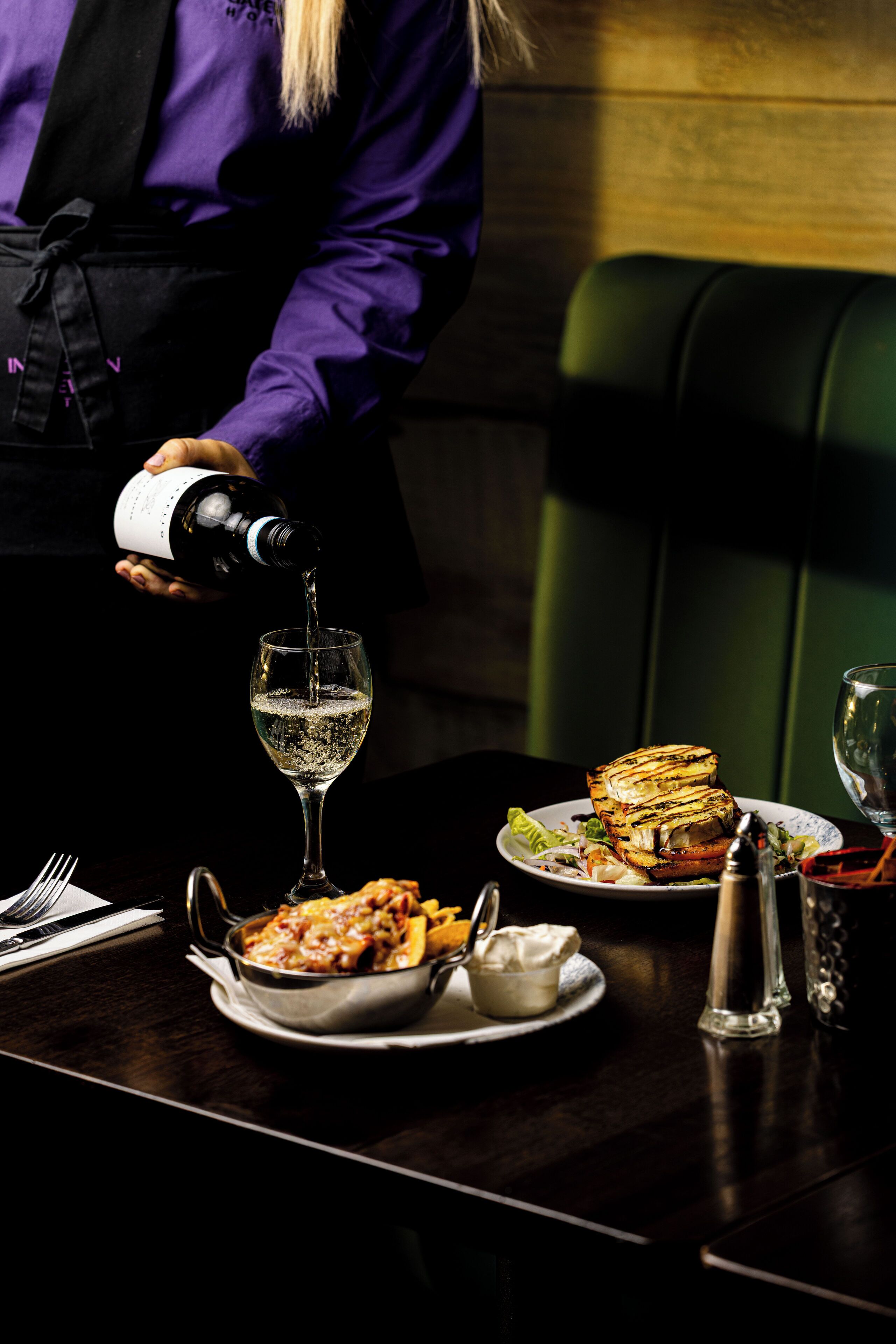 Una persona sirve vino en una copa sobre una mesa con platos de comida y un salero.