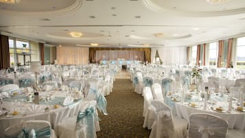 Un salón de banquetes con filas de mesas dispuestas para un evento formal, destacando manteles blancos y sillas con lazos azul claro.