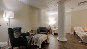 A room with a wooden floor, two armchairs, a small table with a lamp, and a bed with a white bedspread.