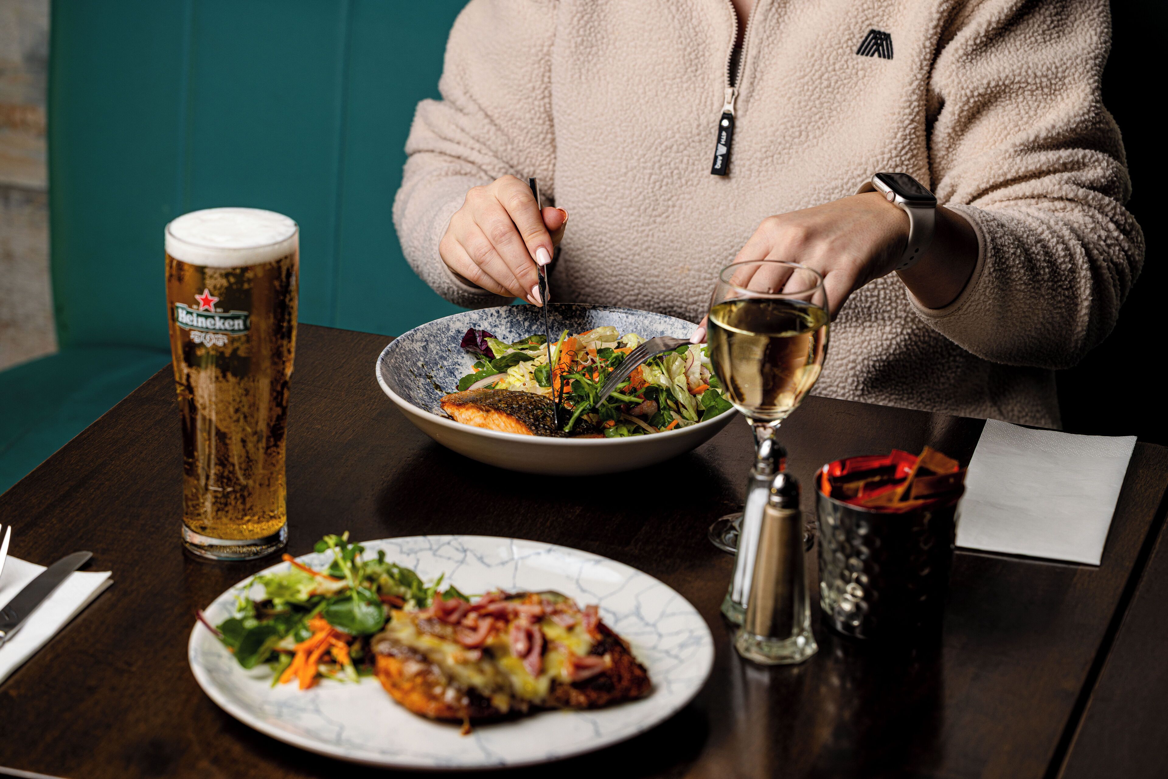 Una persona cenando en una mesa con una copa de cerveza, una copa de vino y un plato de comida.