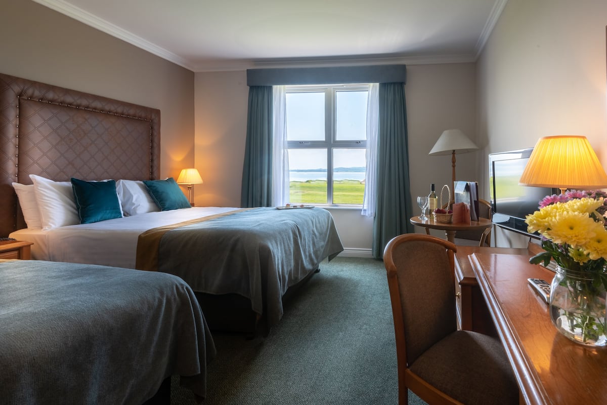 Habitación de hotel con dos camas, un escritorio con lámpara, una silla y vista al mar.