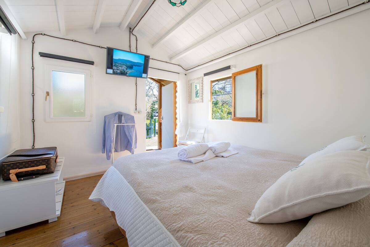 A bedroom with a bed, a television mounted on the wall, a suitcase on a nightstand, and a window with a view of greenery.