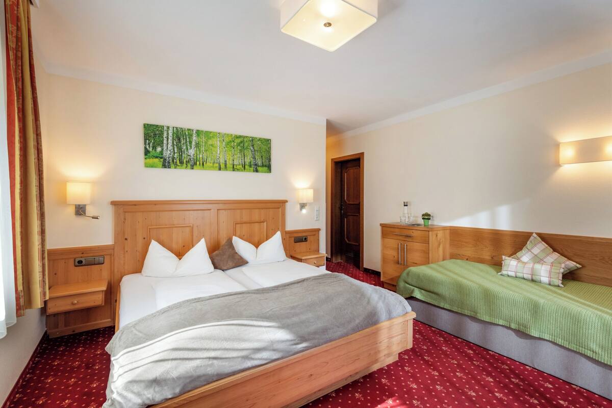 Hotelkamer met een groot bed, een kleiner bed, een houten hoofdbord, een nachtkastje, een lamp, een schilderij aan de muur en een roodgekleurd tapijt.