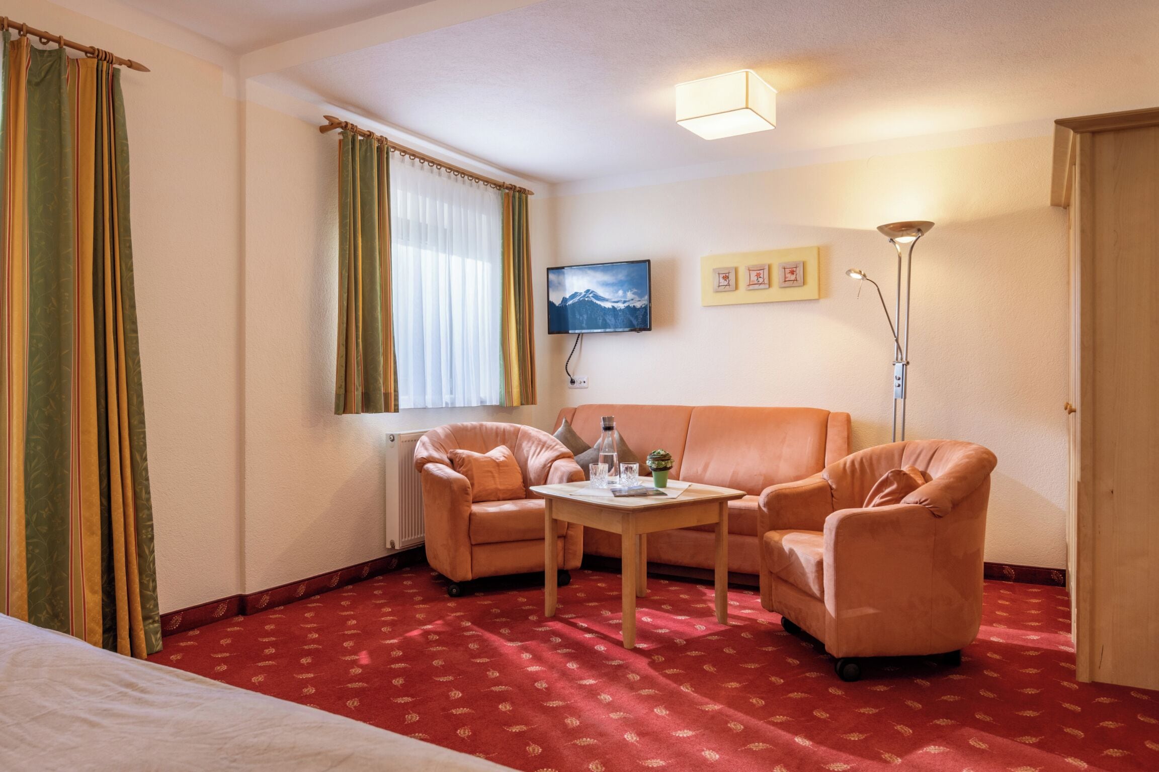 A hotel room with a red carpet, two leather armchairs, a wooden coffee table, a bed, a flat-screen TV, and a floor lamp.