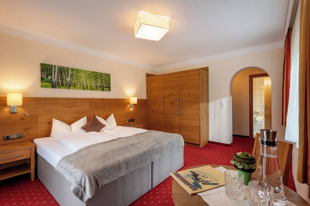 Hotelkamer met een bed, een houten garderobe, bedlampjes, een schilderij aan de muur en een klein tafeltje met een boek en glazen.