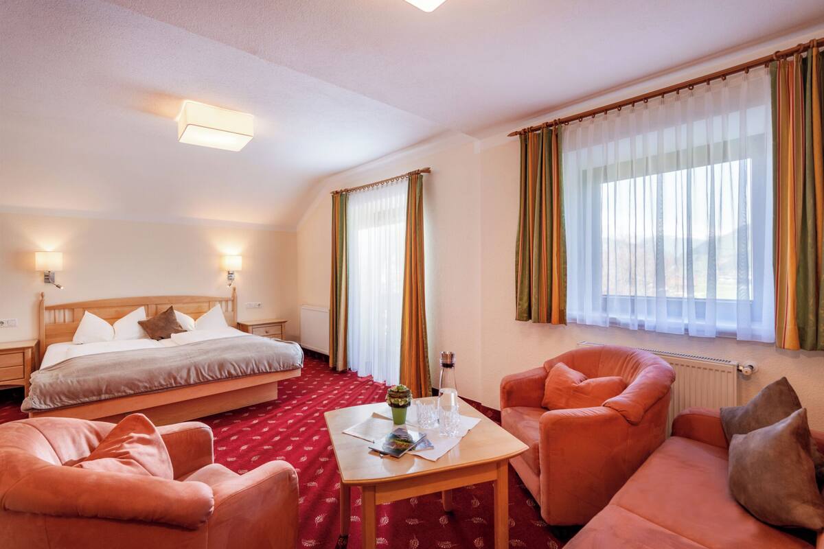 Een hotelkamer met een bed, twee fauteuils, een tafel met een boek en een glas, en gordijnen voor de ramen.