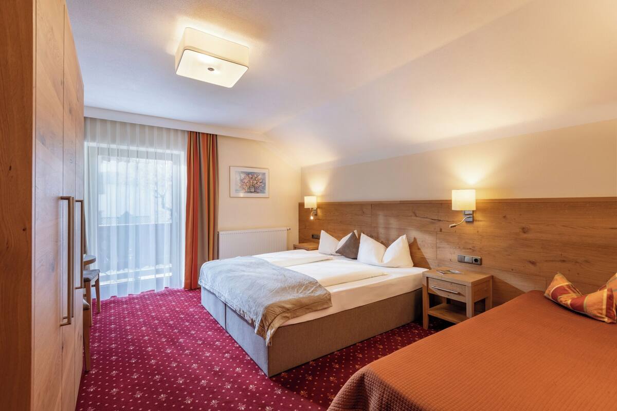 Hotelkamer met een groot bed, nachtkastje, houten hoofdeinde, raam met gordijnen en een rood tapijt.