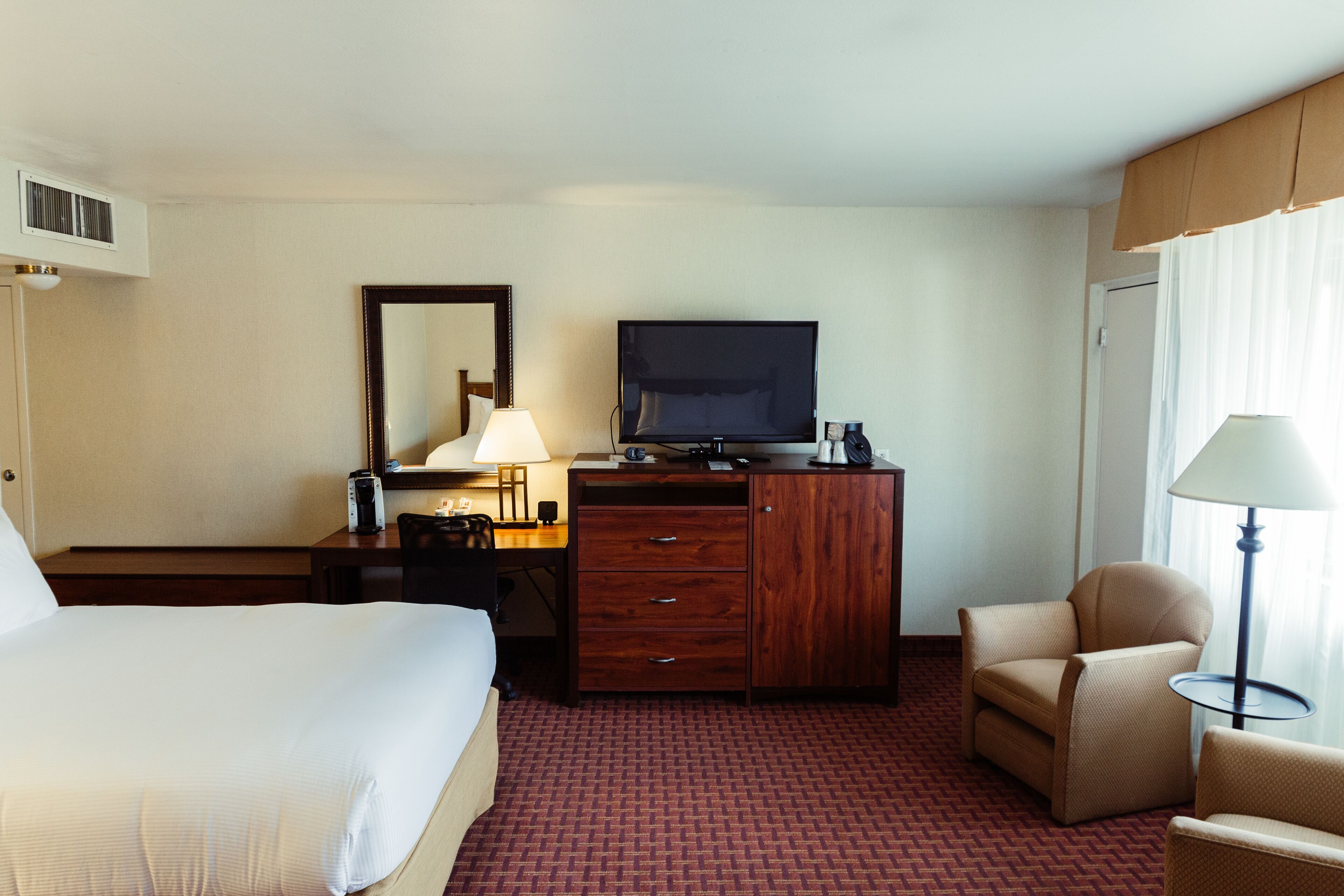 A hotel room with a bed, a desk with a lamp, a television, a chair, and a wardrobe.