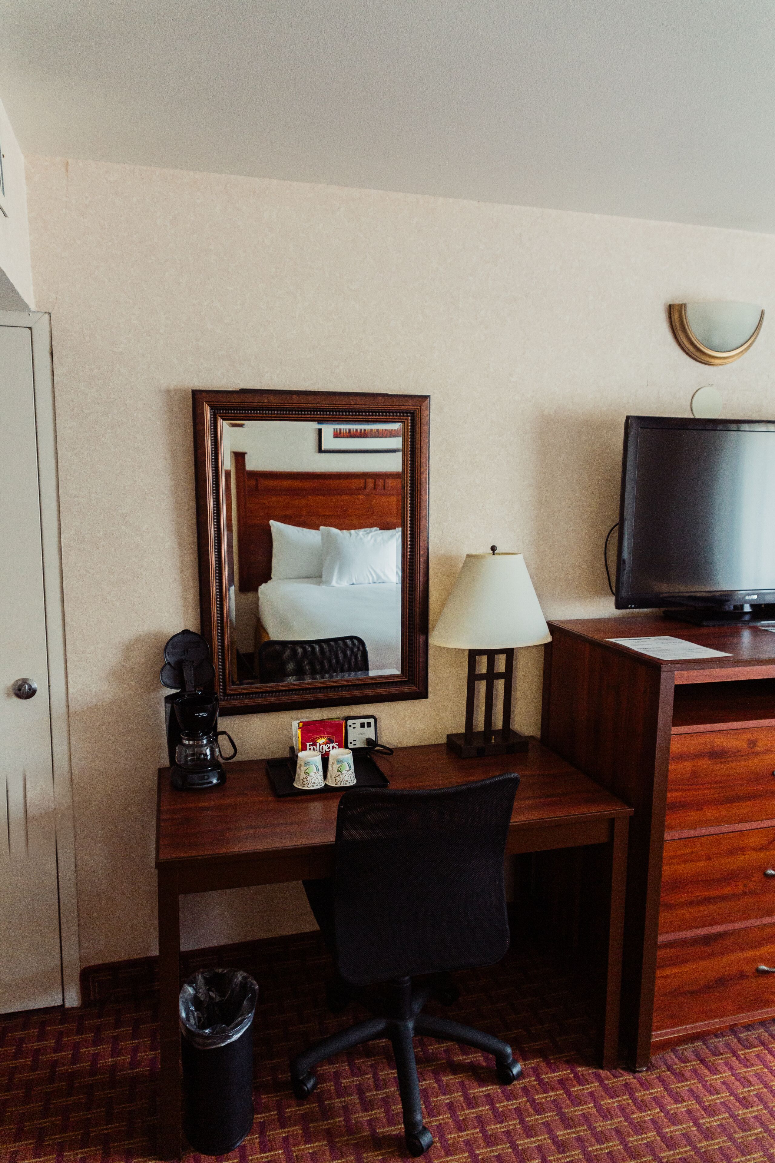 A hotel room with a wooden desk, a black office chair, a television, a lamp, a coffee maker, and a trash can.