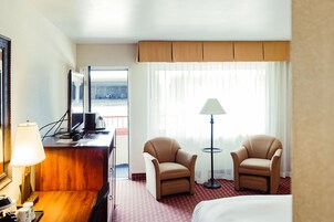 Habitación de hotel con una cama, dos sillones, un escritorio con una lámpara, una computadora y vistas a un paisaje urbano por la ventana.