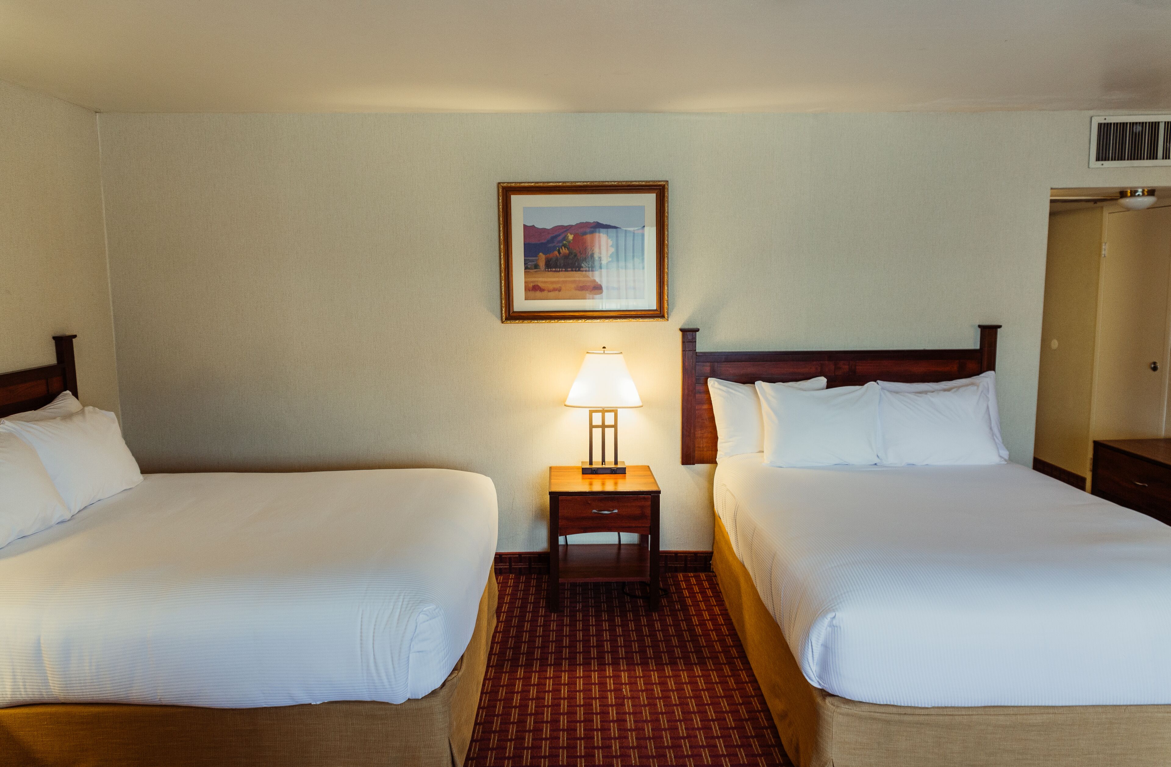 A hotel room with two single beds, a nightstand with a lamp, a framed picture on the wall, and a carpeted floor.