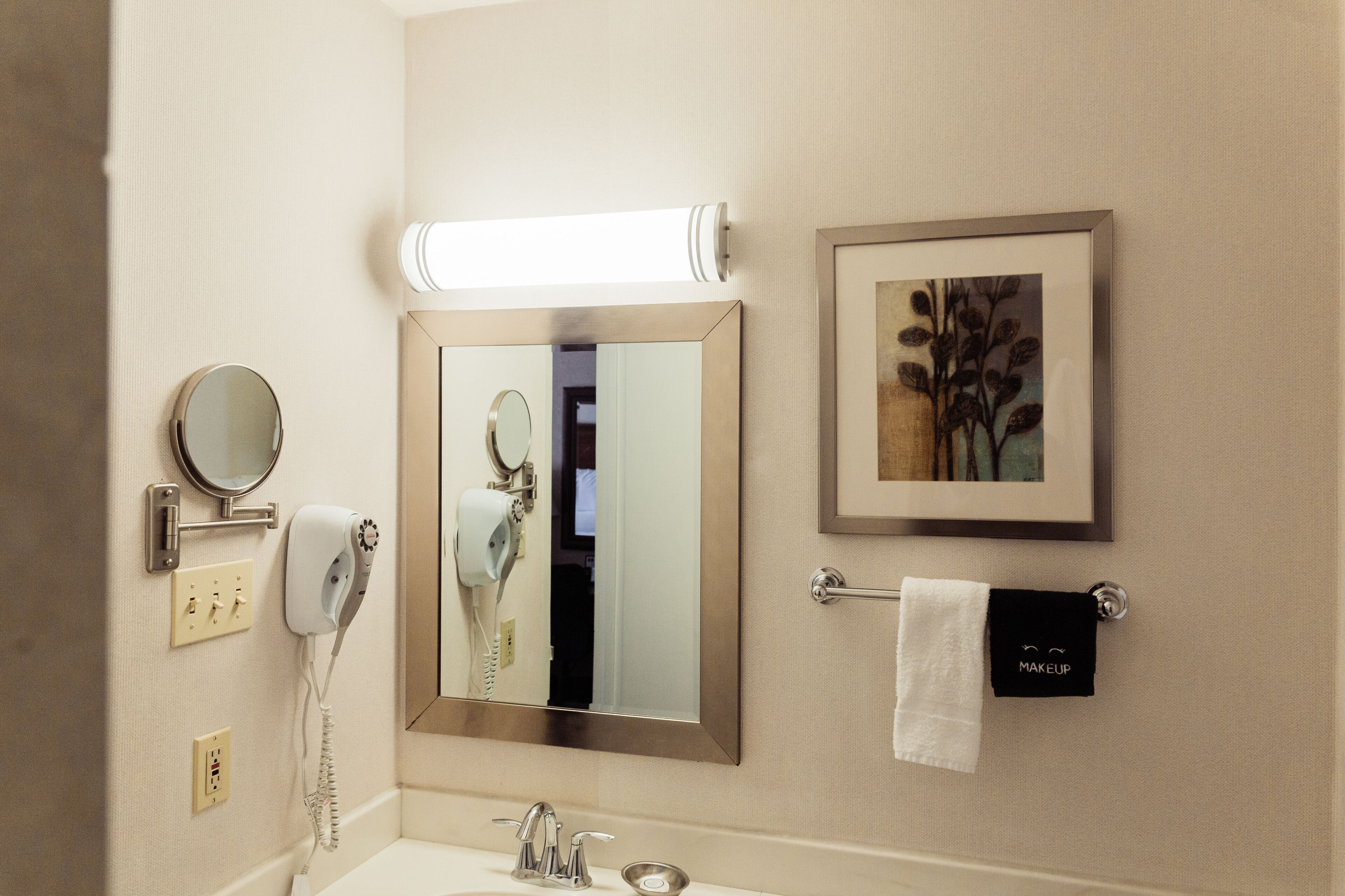 A bathroom with a large mirror, a hairdryer, a telephone, and a framed picture on the wall.