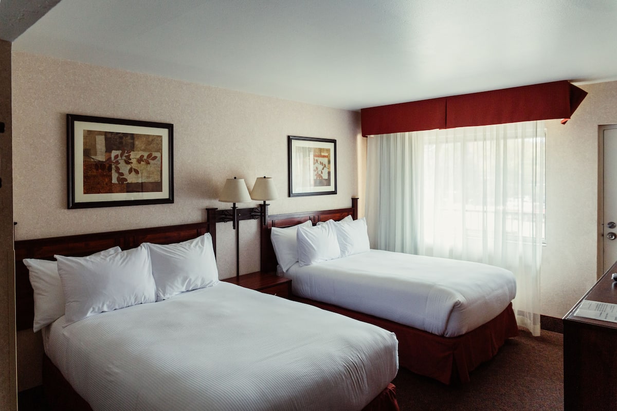A hotel room with two single beds, a nightstand with a lamp, a framed picture on the wall, and a window with sheer curtains.