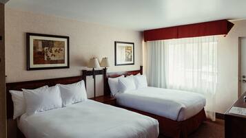 Una habitación de hotel con dos camas individuales, una mesita de noche con lámpara, un cuadro en la pared y una ventana con cortinas translúcidas.