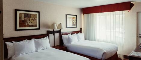 A hotel room with two single beds, a nightstand with a lamp, a framed picture on the wall, and a window with sheer curtains.